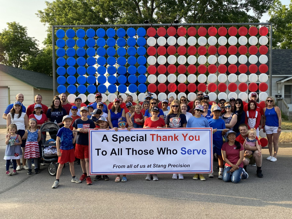 A group gathered in front of Stang Precision’s frisbee flag with a banner thanking all those who serve